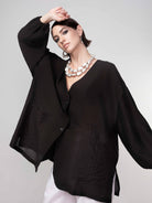 Woman wearing a black lightweight blouse with a relaxed fit, wide sleeves, and button-front detail, paired with white pants and statement silver jewellery against a neutral background.