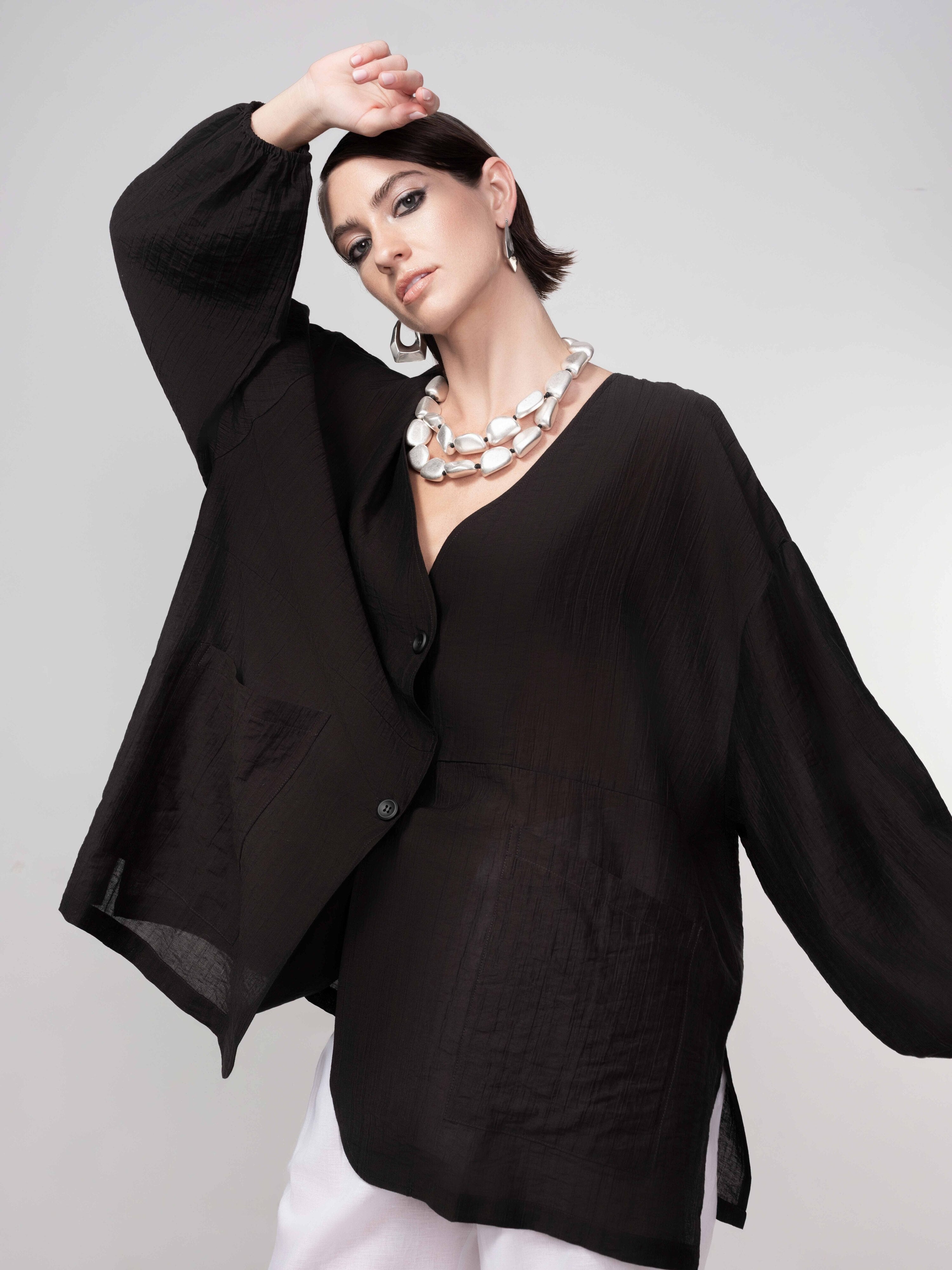 Woman wearing a black lightweight blouse with a relaxed fit, wide sleeves, and button-front detail, paired with white pants and statement silver jewellery against a neutral background.