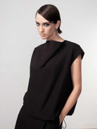 Woman wearing a black relaxed cocoon-style pullover top with cap sleeves and a high neckline, shown in a close-up studio shot with hands in pockets.