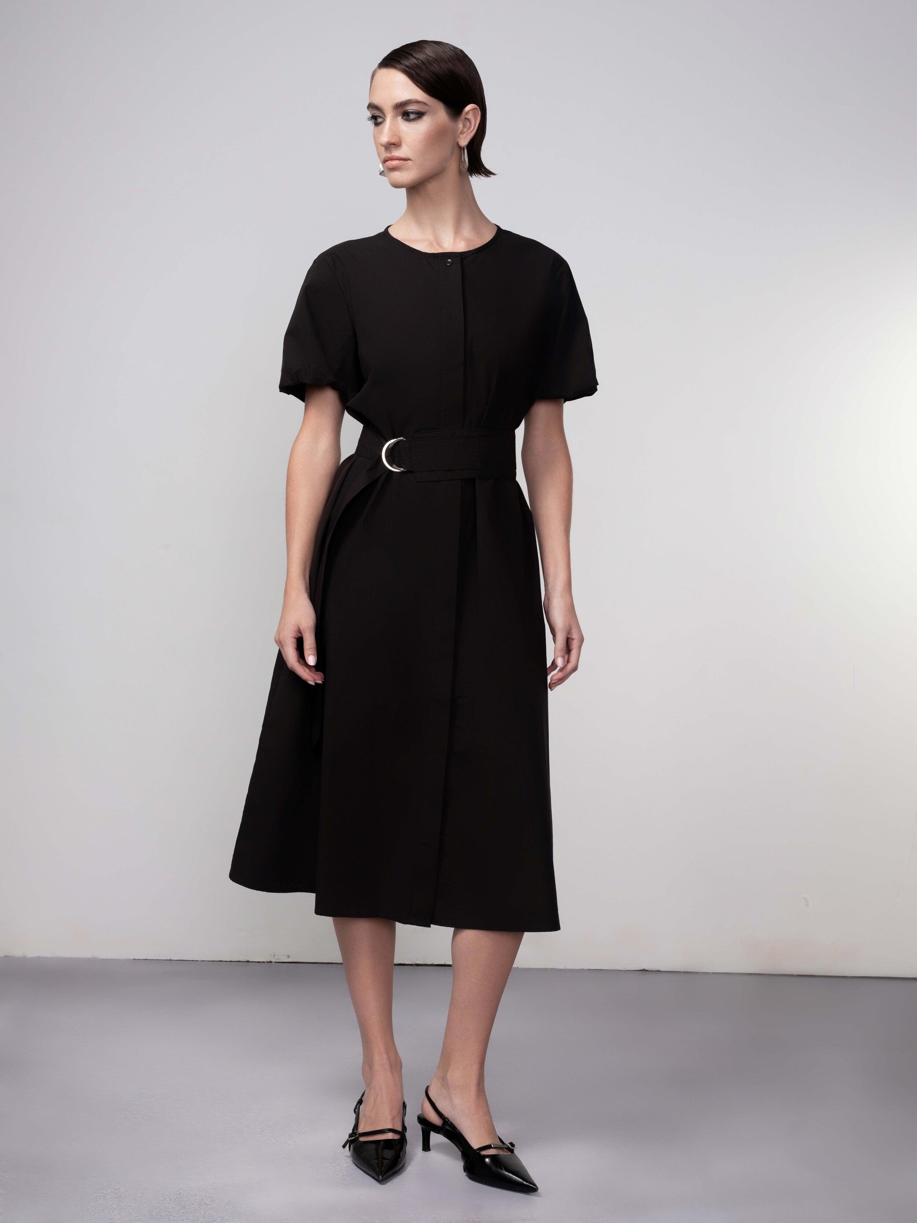 Woman wearing a black cotton midi dress with short sleeves, a round neckline, and a belted waist, paired with pointed-toe heels, standing against a neutral background.