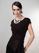 Woman wearing a black drape-knot top with cap sleeves, styled with wide-leg pants and a statement necklace, front-facing pose.