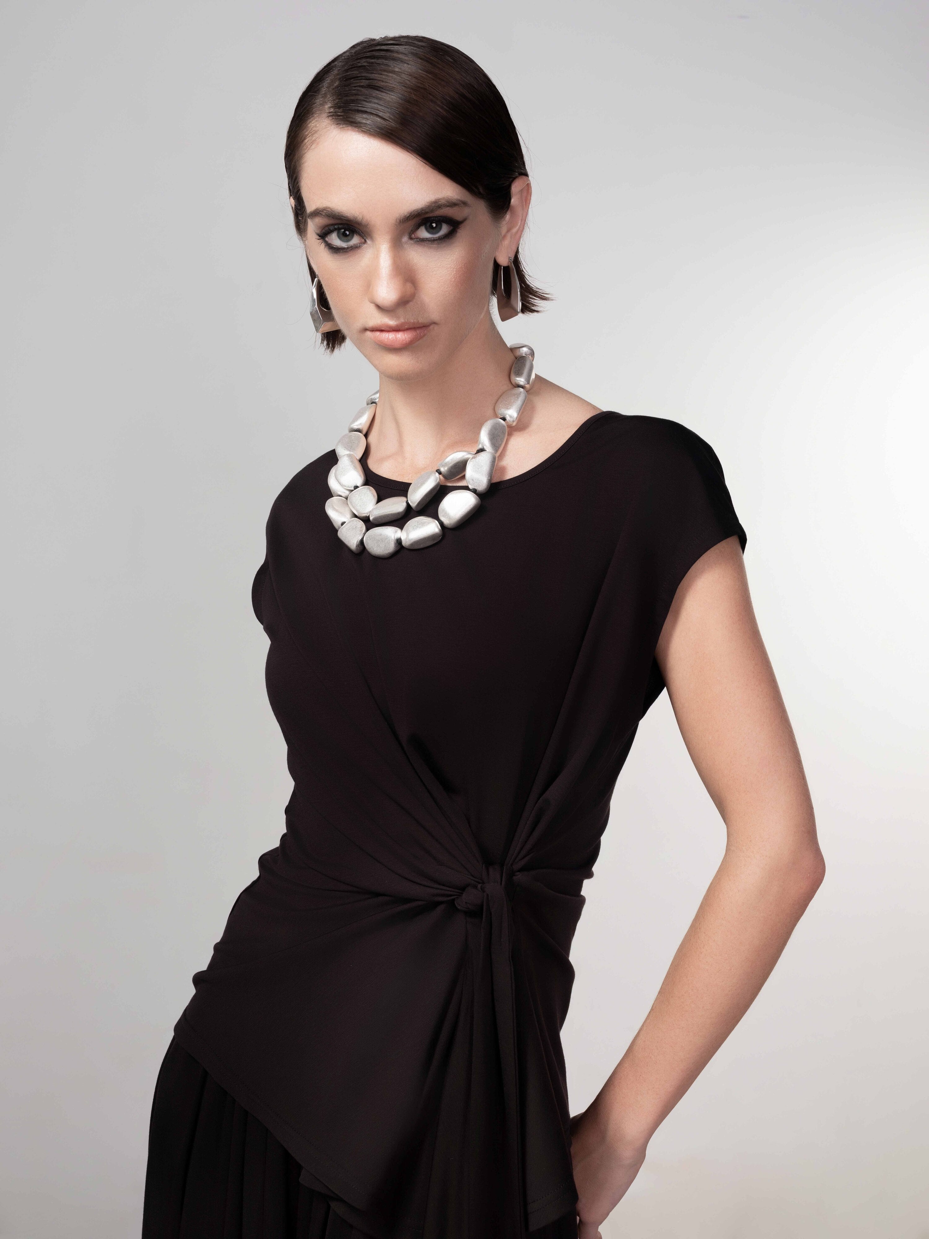 Woman wearing a black drape-knot top with cap sleeves, styled with wide-leg pants and a statement necklace, front-facing pose.