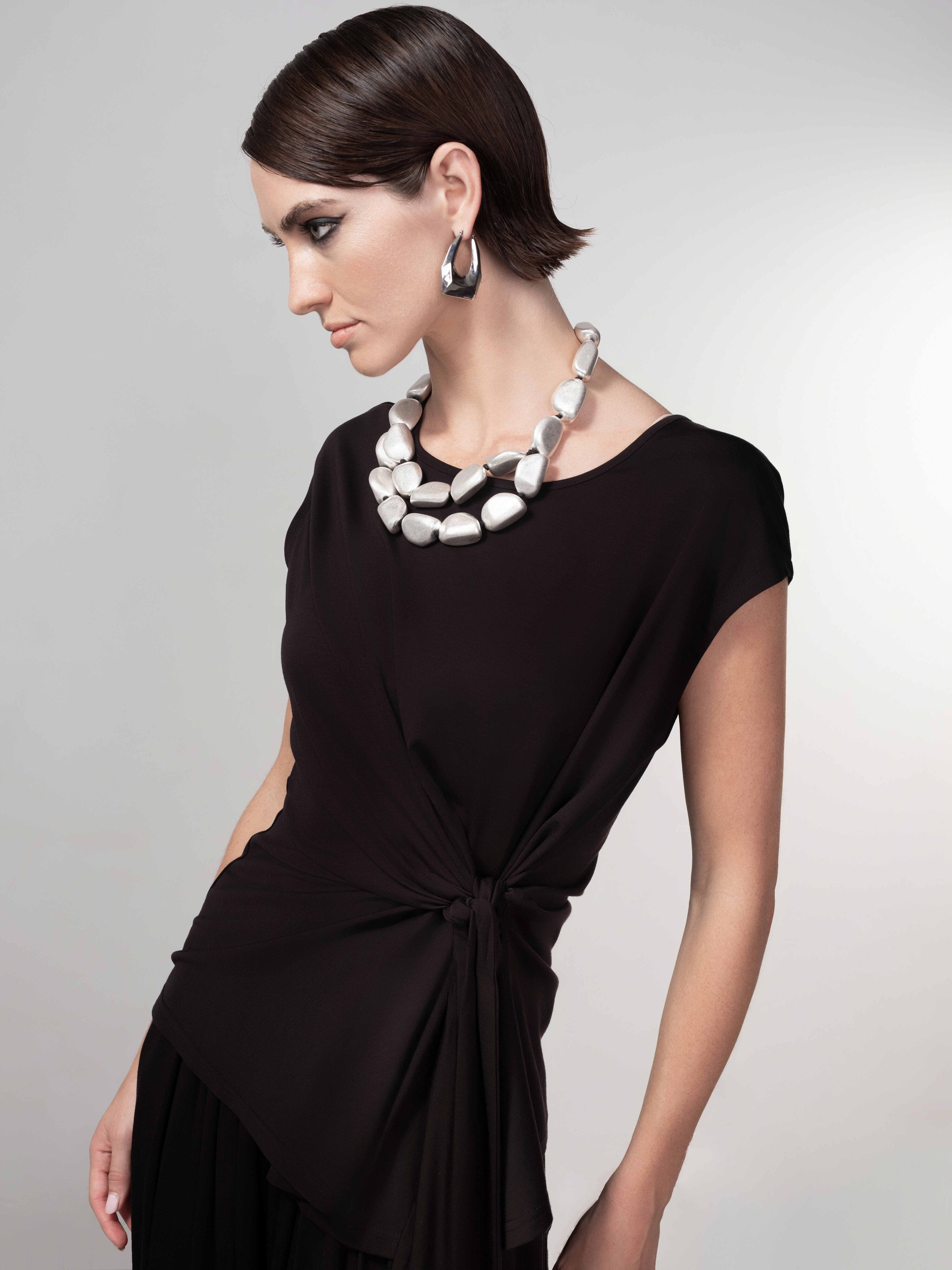 Side profile of a woman in a black draped pullover top with a side-knot detail, paired with wide-leg pants and bold jewellery.