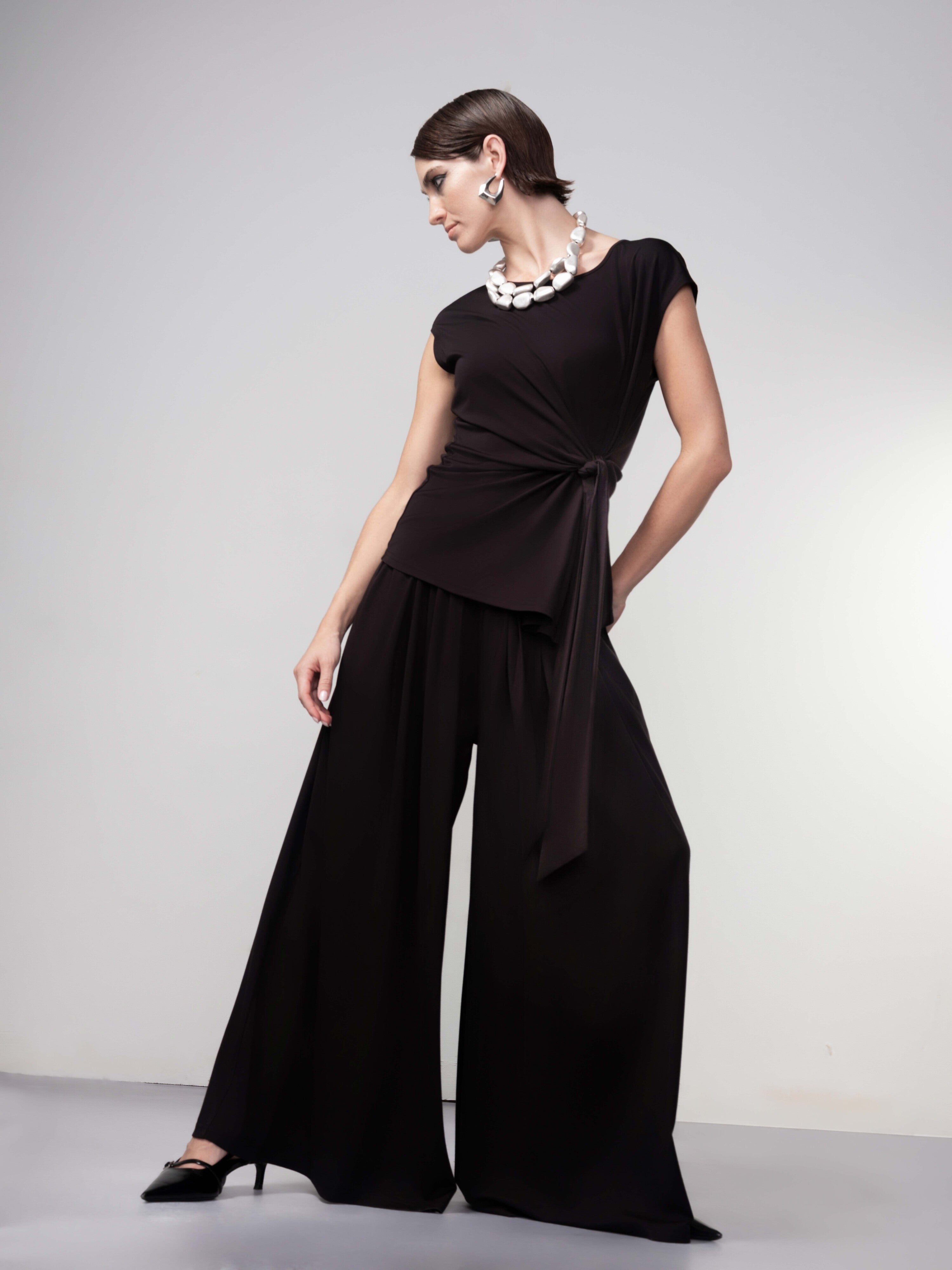 Full-length view of a woman wearing a black drape-knot top with matching wide-leg pants and heels, styled with a chunky necklace and earrings.