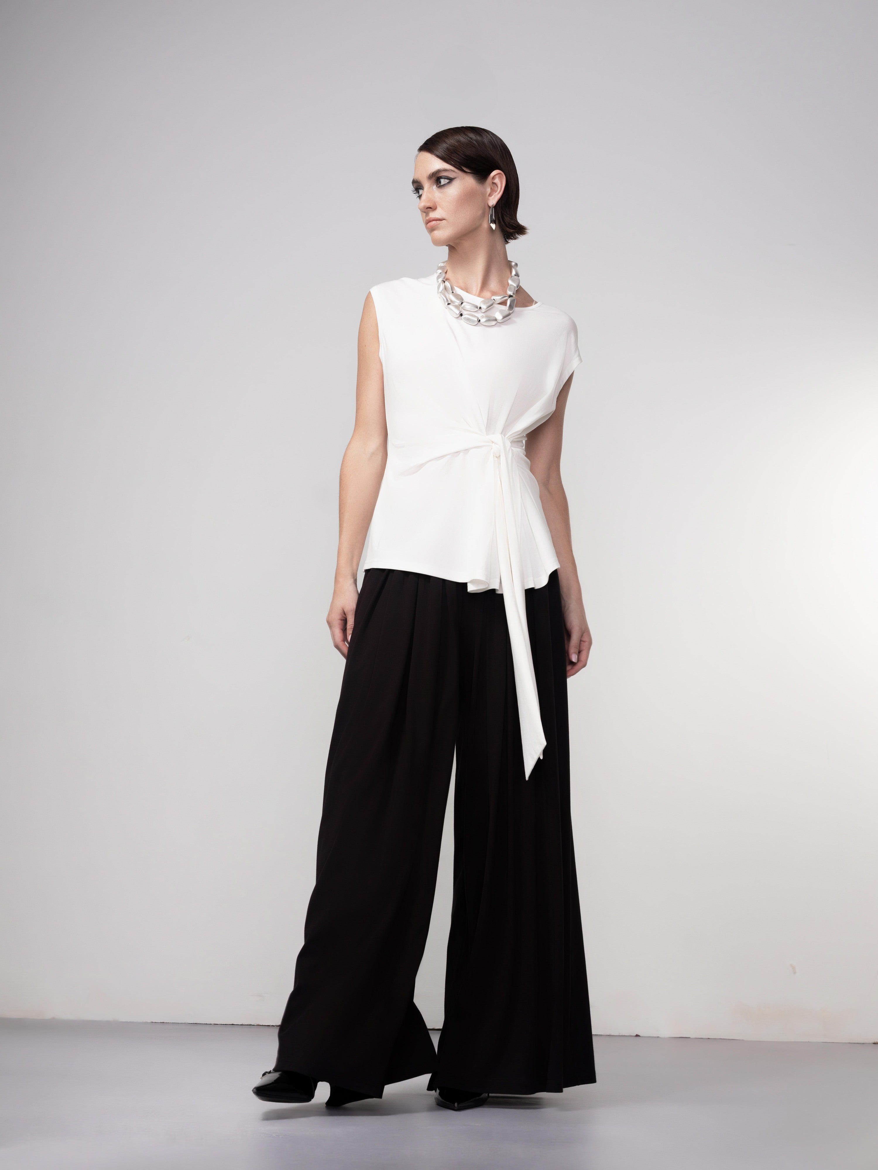 Model wearing a sleeveless white draped blouse with a side tie detail paired with black wide-leg pants and black shoes, styled with a chunky necklace against a neutral studio background.