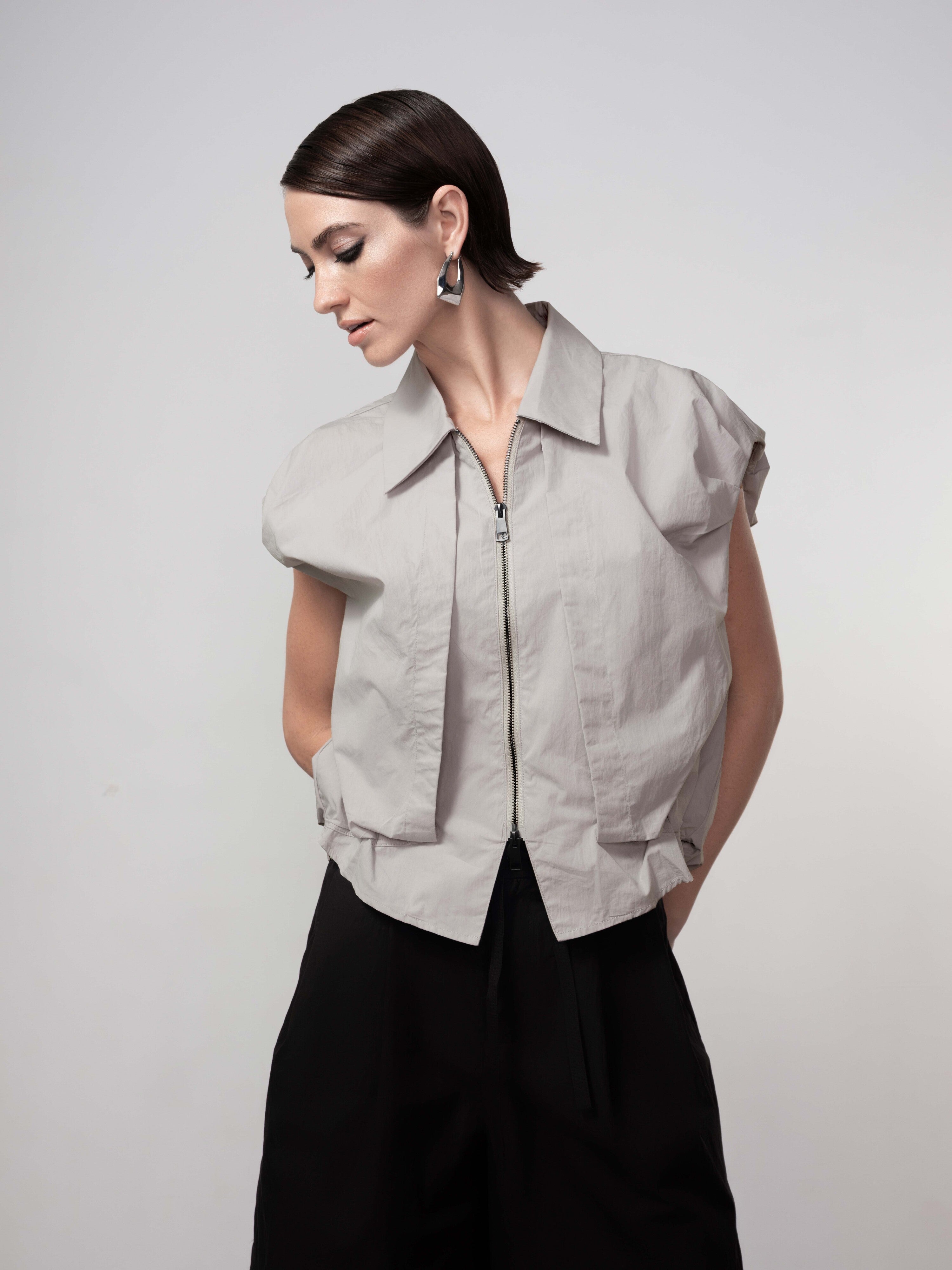 Woman wearing a silver zip-front vest cardigan with cap sleeves, paired with wide-leg trousers, looking downward in a studio setting.