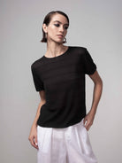 Woman wearing a black lightweight sheer knit short-sleeve sweater with a relaxed fit, paired with white wide-leg pants, shown in a studio setting.