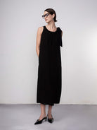 Woman wearing a black sleeveless midi dress with gathered neckline and tie-shoulder detail, paired with pointed-toe flats and glasses.