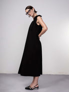 Woman wearing a black sleeveless midi dress with gathered neckline and tie-shoulder detail, paired with pointed-toe flats and glasses.