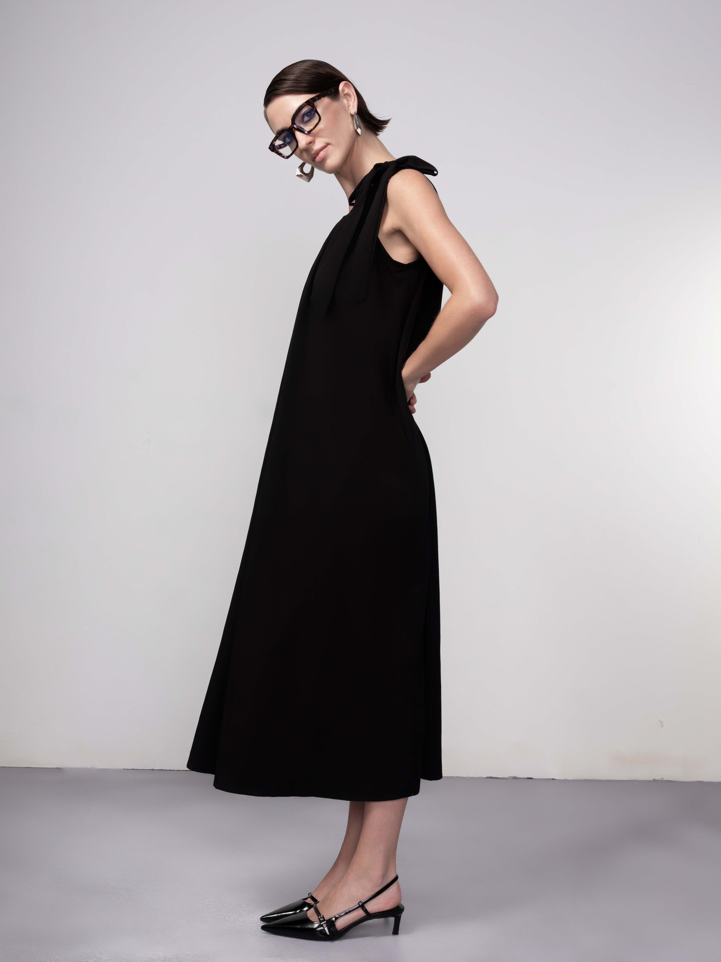 Woman wearing a black sleeveless midi dress with gathered neckline and tie-shoulder detail, paired with pointed-toe flats and glasses.
