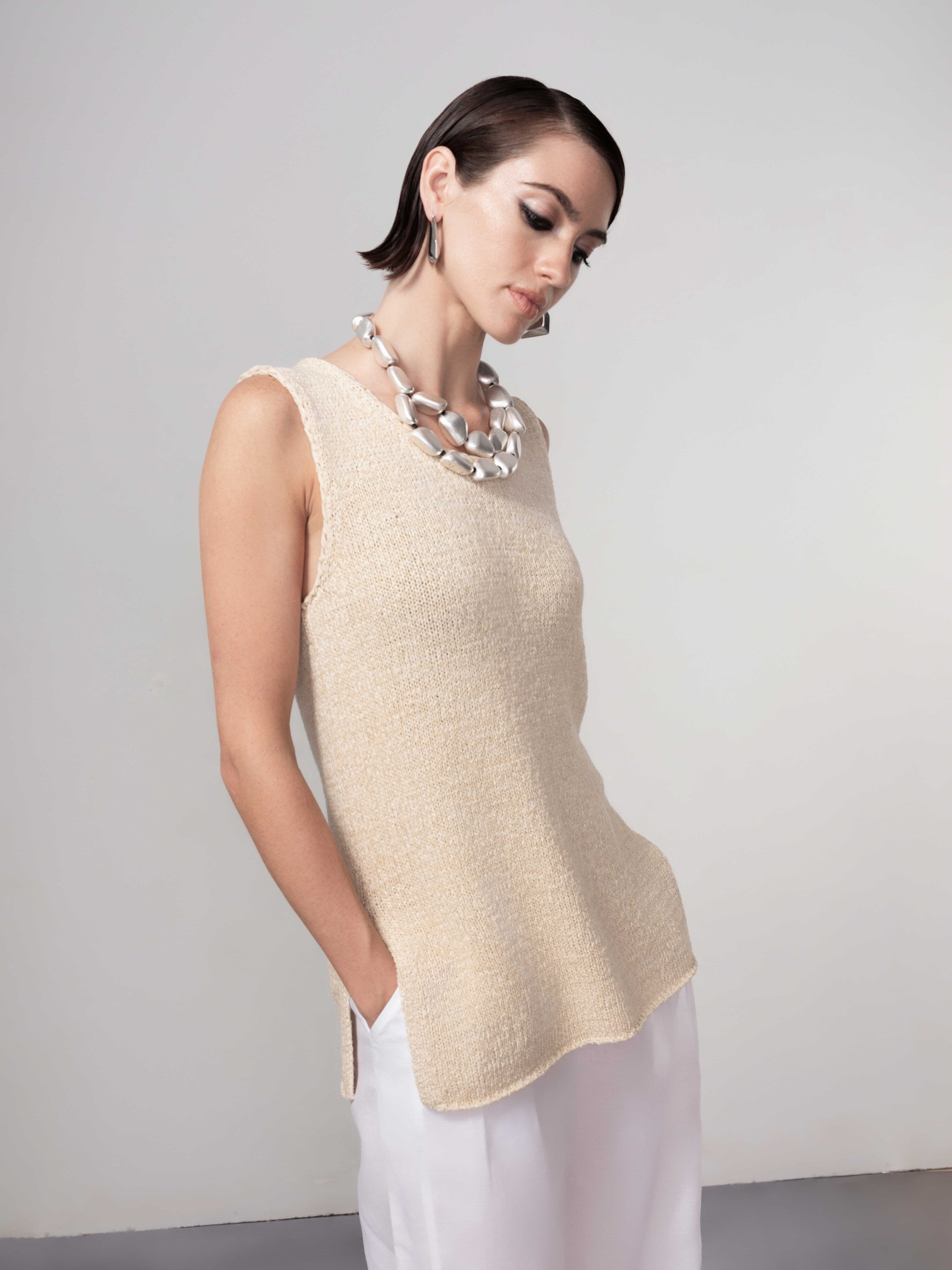 Side view of a woman wearing a sleeveless beige textured knit shell top with side slits, styled with light trousers and a statement necklace.
