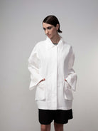 Model wearing a white oversized utility jacket with wide sleeves and front pockets, paired with black shorts against a neutral background.