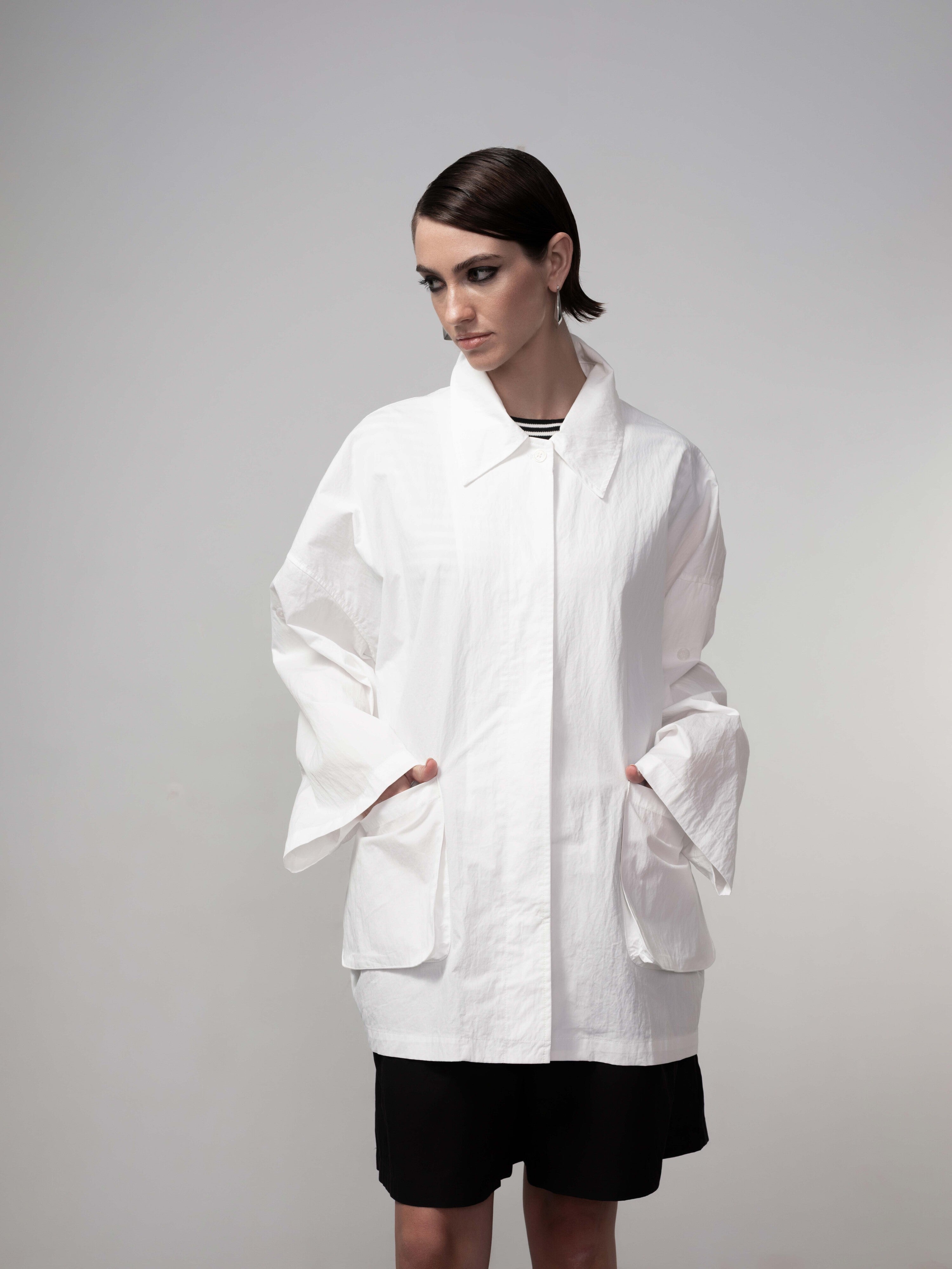 Model wearing a white oversized utility jacket with wide sleeves and front pockets, paired with black shorts against a neutral background.