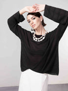 Woman wearing a black lightweight knit sweater with long sleeves, styled with a white skirt, statement necklace, and geometric earrings, posing with arms raised.