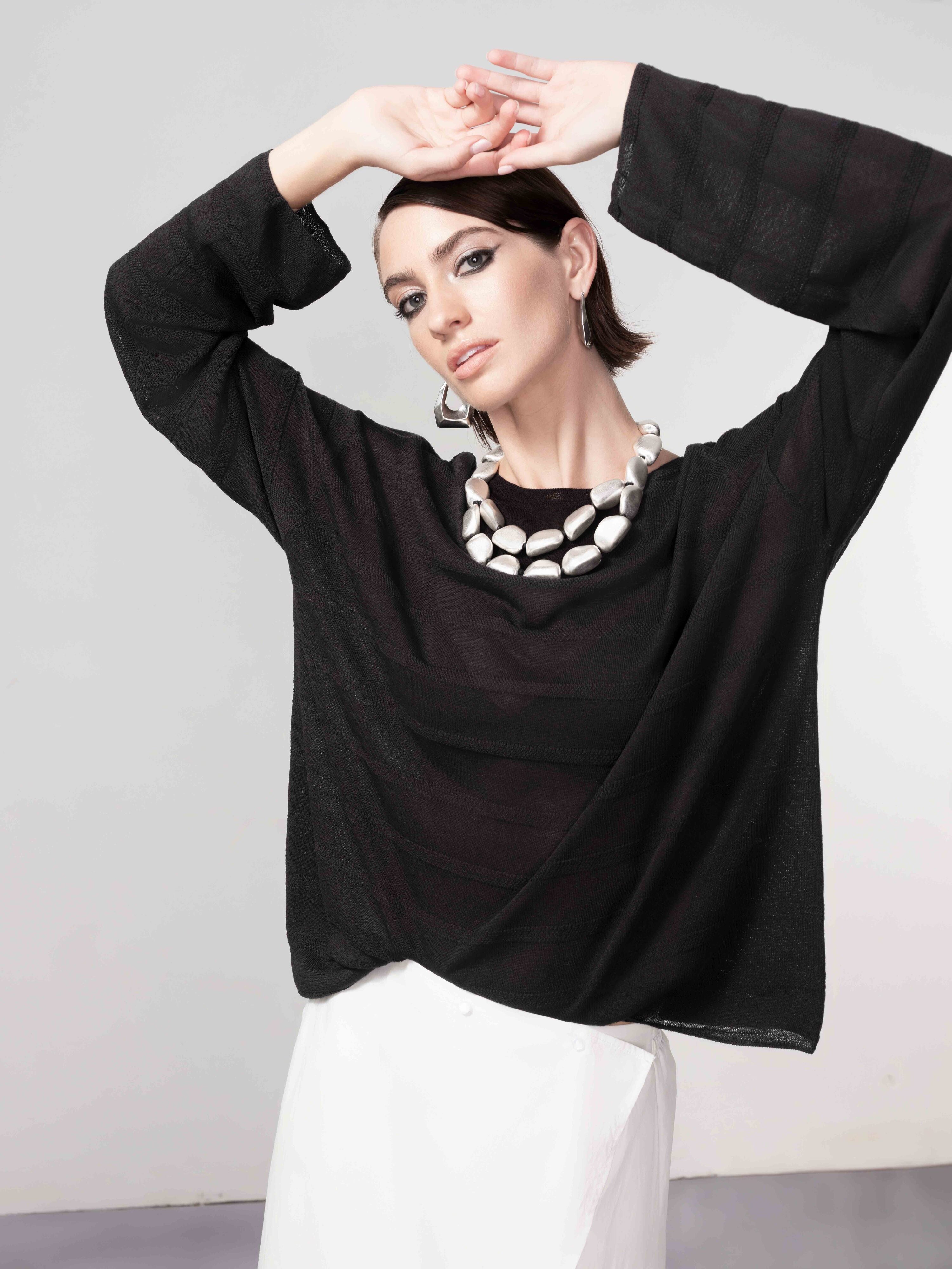 Woman wearing a black lightweight knit sweater with long sleeves, styled with a white skirt, statement necklace, and geometric earrings, posing with arms raised.