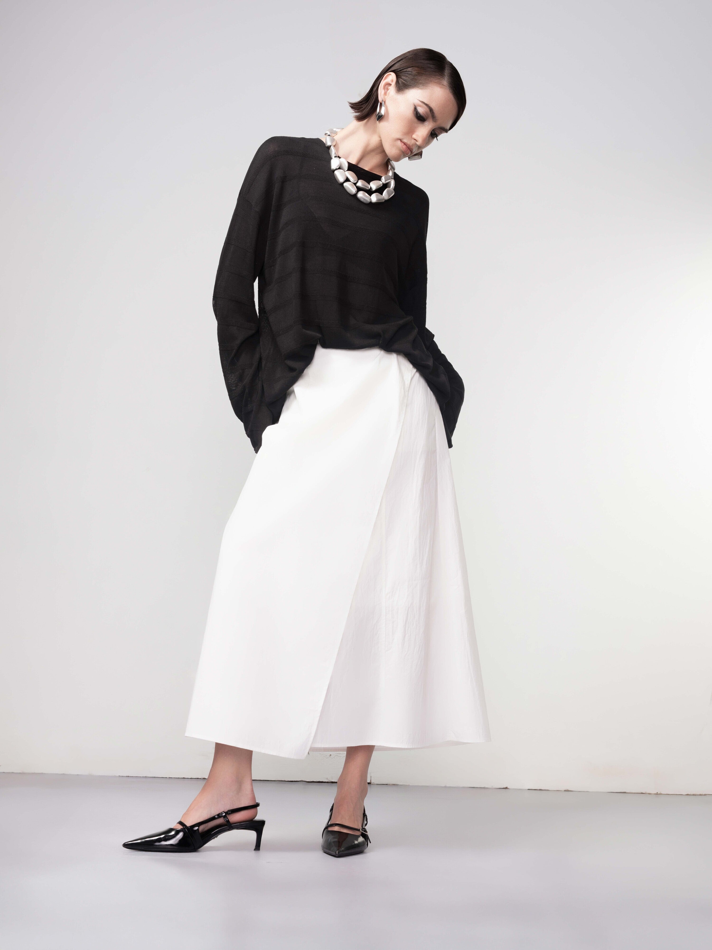 Full-length view of a woman in a black relaxed-fit knit sweater paired with a white midi skirt and black pointed slingback heels.