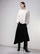 Woman wearing a soft white lightweight knit sweater styled with a black midi skirt and black knee-high heeled boots, hands in pockets.