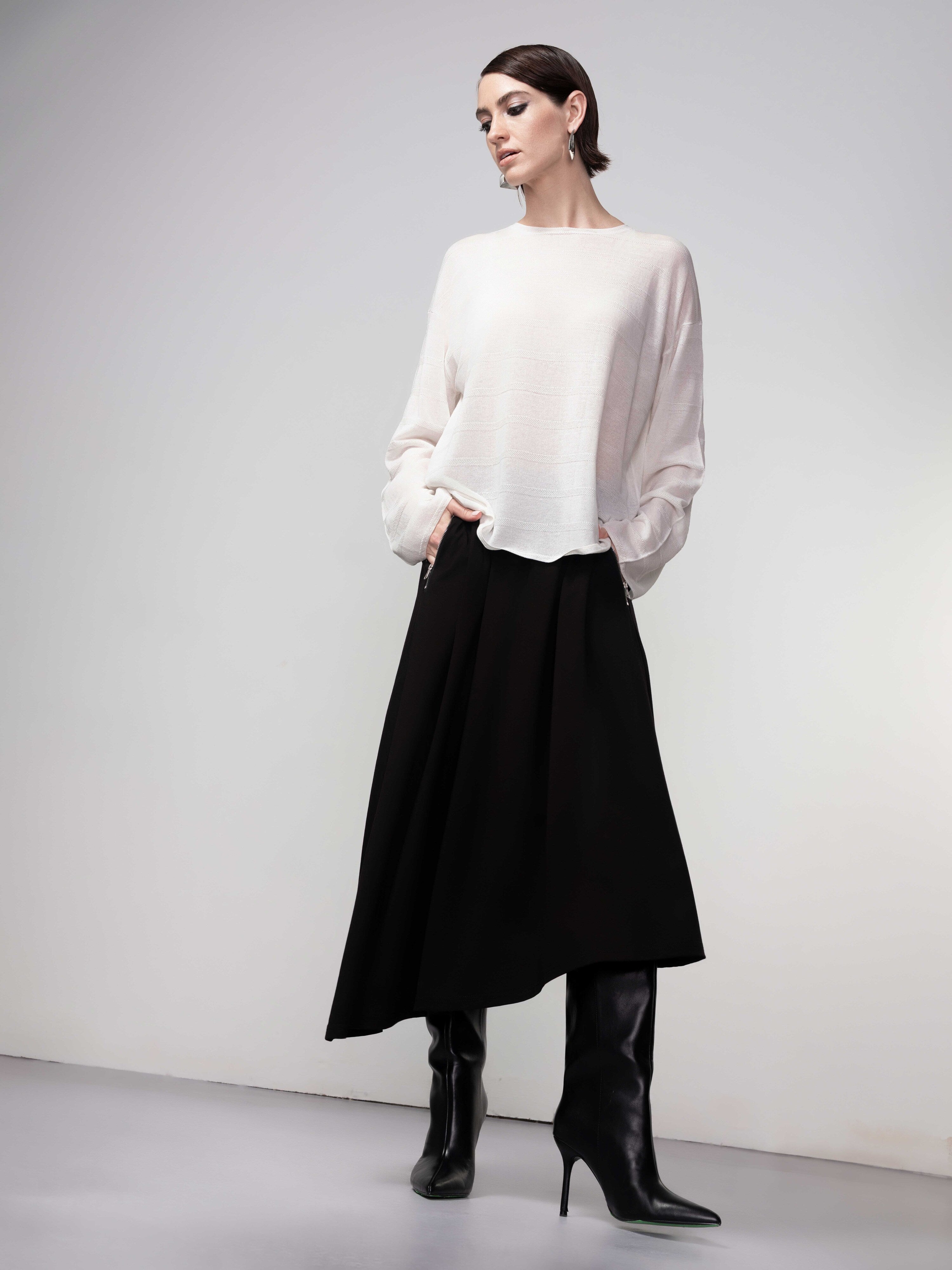 Woman wearing a soft white lightweight knit sweater styled with a black midi skirt and black knee-high heeled boots, hands in pockets.