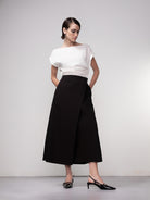 Model wearing a black sleeveless top paired with a white midi A-line wrap skirt and pointed slingback heels, front view.