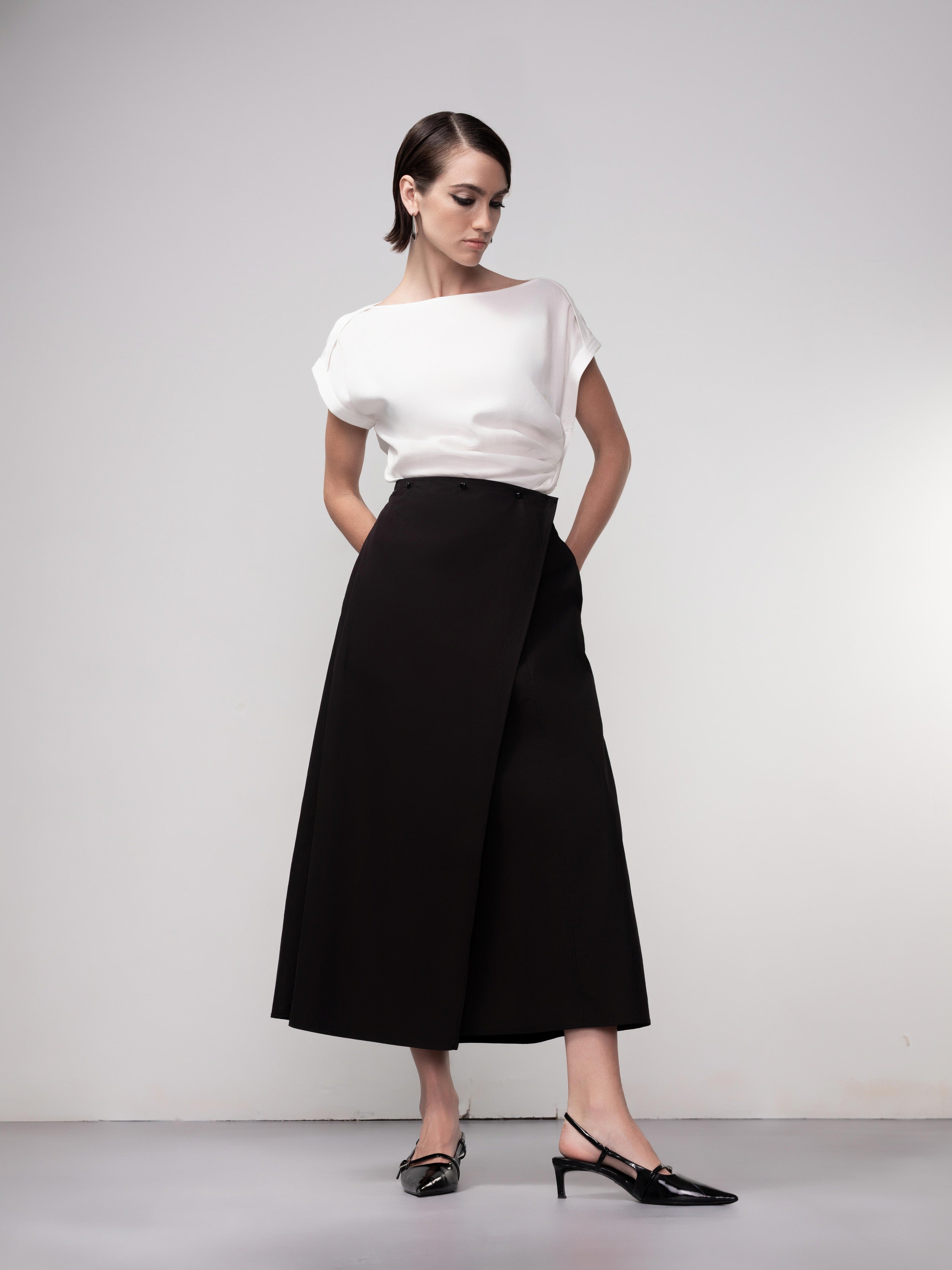 Model wearing a black sleeveless top paired with a white midi A-line wrap skirt and pointed slingback heels, front view.