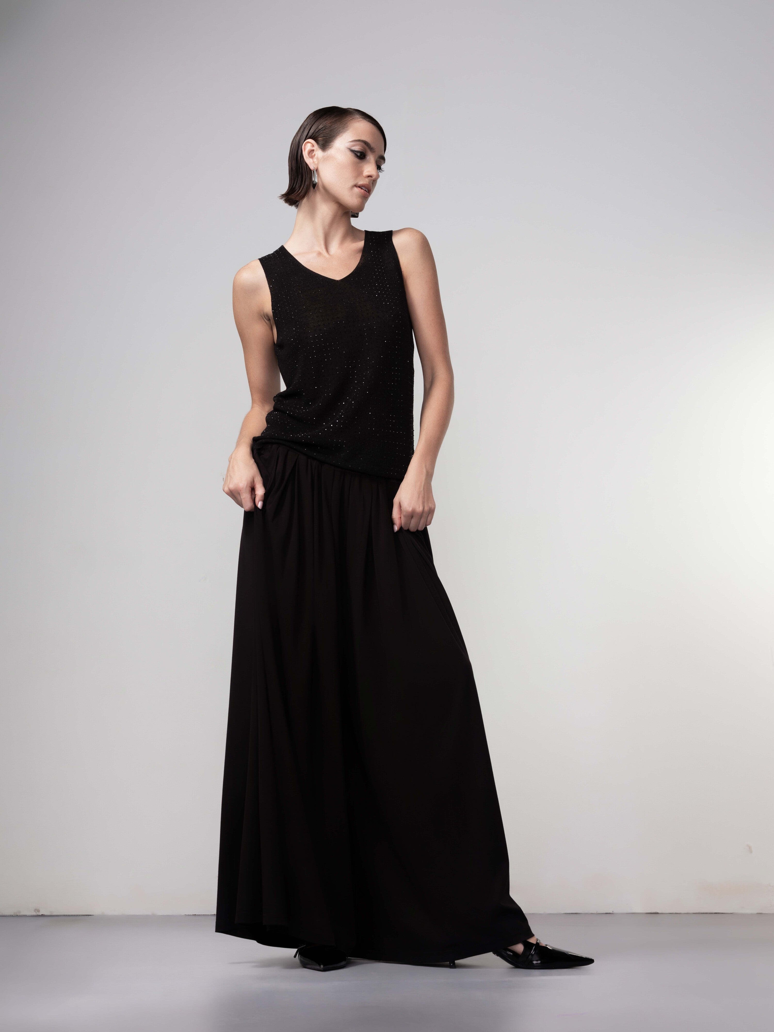 Model wearing a black sleeveless knit tank top paired with a long flowing black skirt and black pointed shoes, standing against a neutral background.