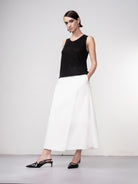 Model wearing a black sleeveless knit tank top styled with a mid-length white skirt and black slingback heels, posing against a neutral studio backdrop.