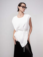 Woman wearing a minimalist sleeveless white blouse with a soft drape and tie waist detail, paired with a black skirt and oversized tortoiseshell glasses, styled in a clean studio setting.