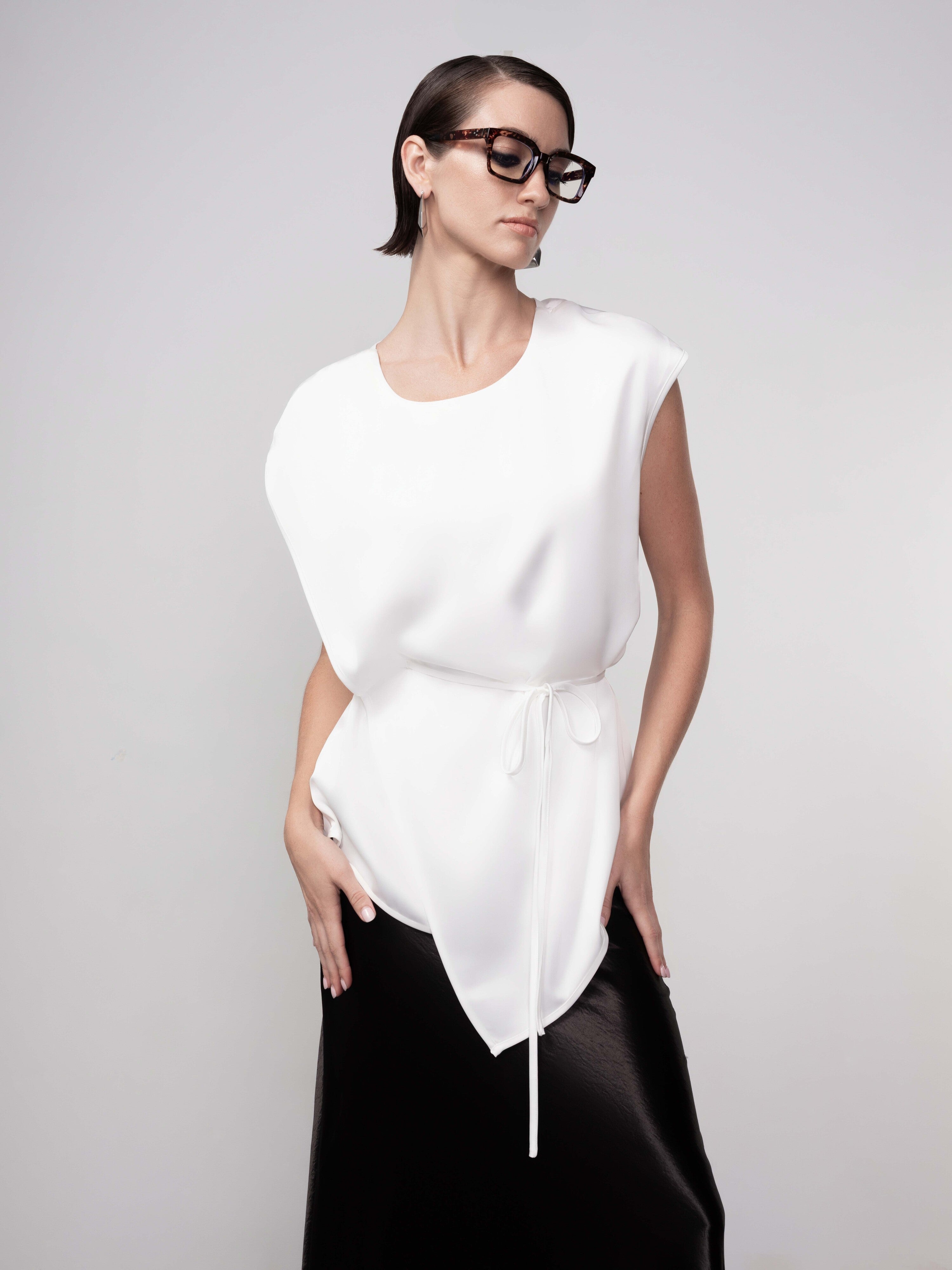 Woman wearing a minimalist sleeveless white blouse with a soft drape and tie waist detail, paired with a black skirt and oversized tortoiseshell glasses, styled in a clean studio setting.