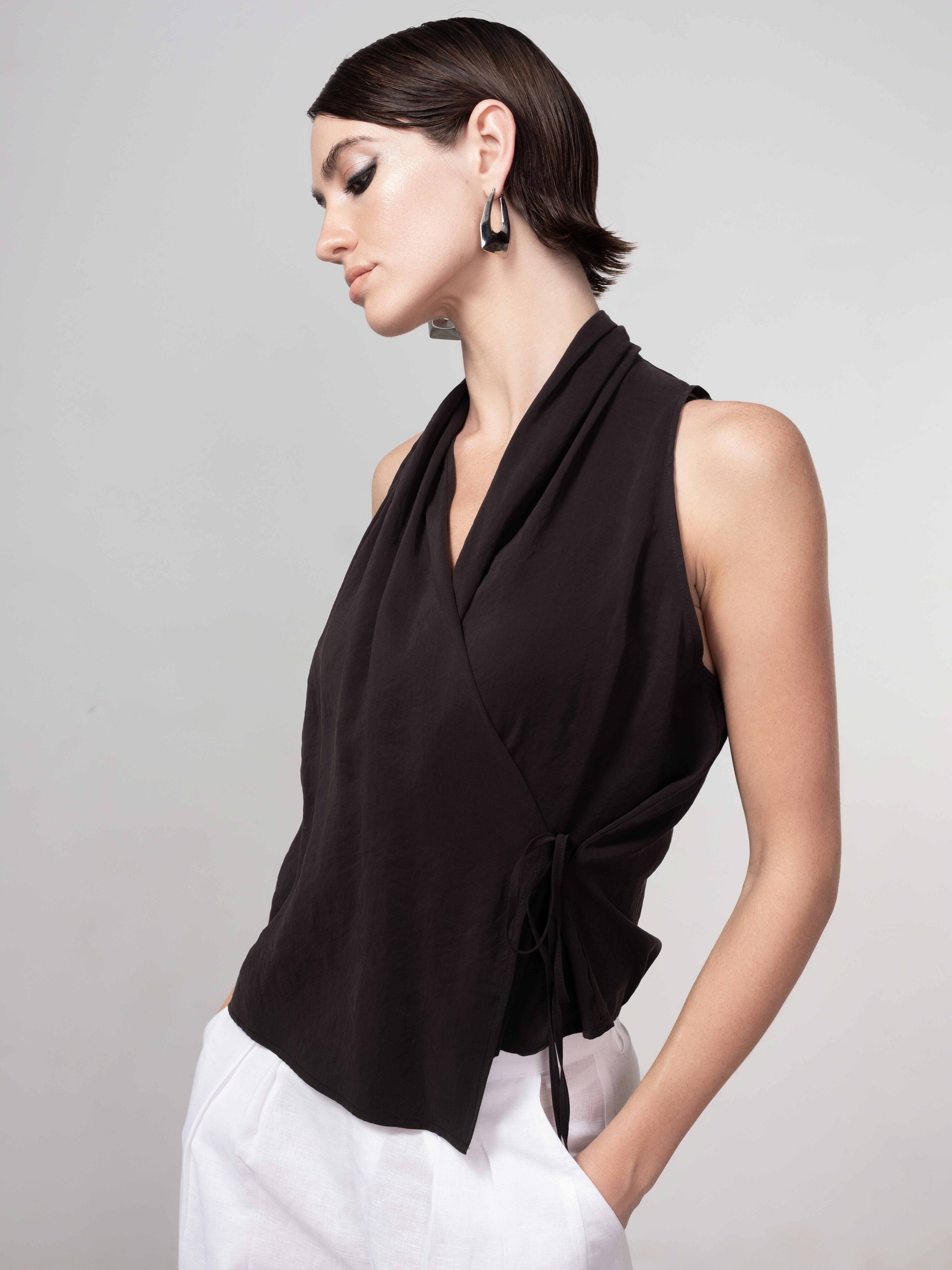 Woman wearing a black sleeveless wrap blouse with a soft drape and side tie, styled with light trousers