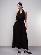 Woman wearing a black halter-neck midi dress with a relaxed draped silhouette, elasticized waist, and fluid skirt, paired with pointed slingback heels against a neutral studio background.