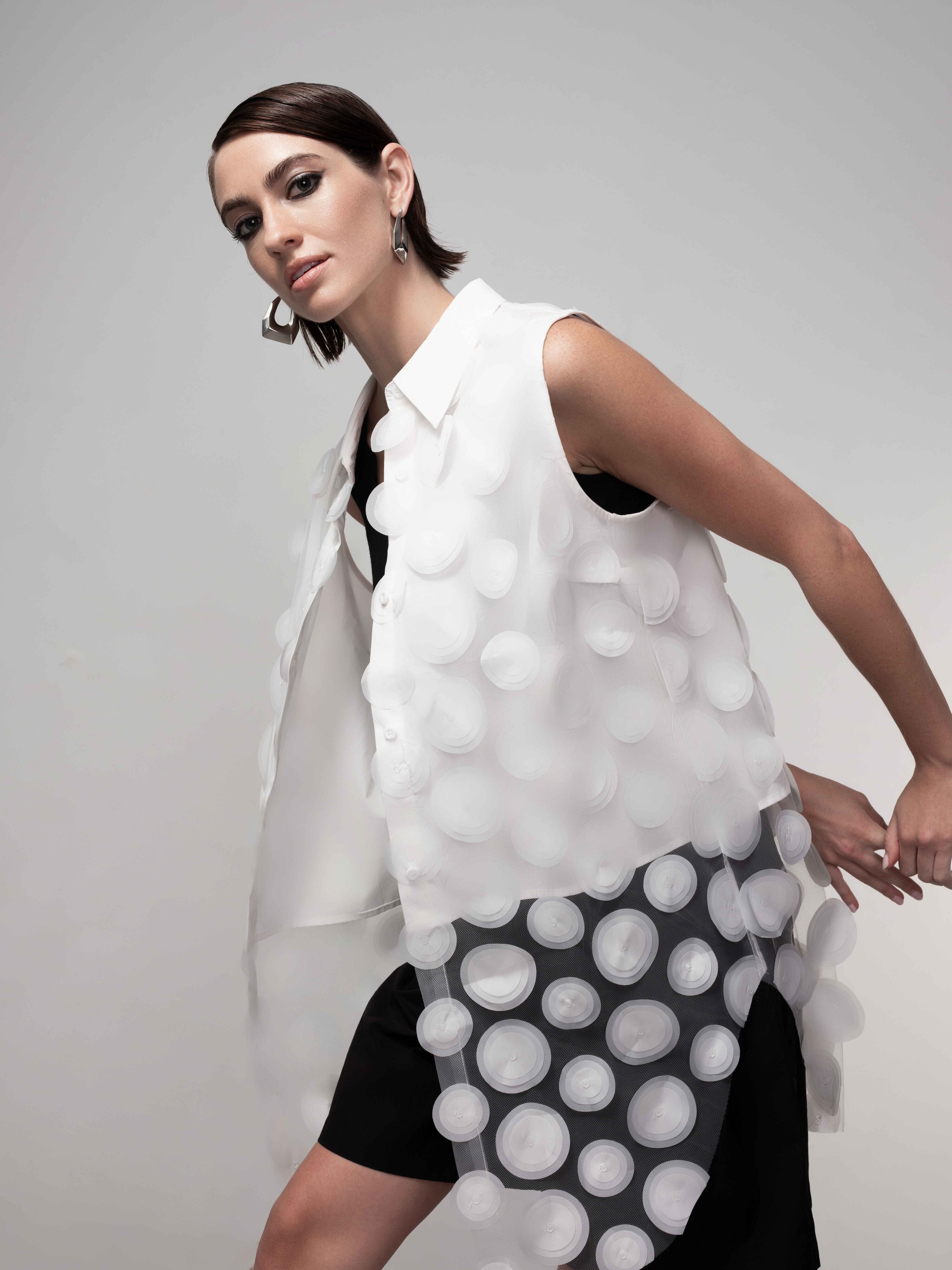 Angled close-up of a sheer white textured tunic vest with circular appliqué over a black dress, highlighting the lightweight fabric and layered design.