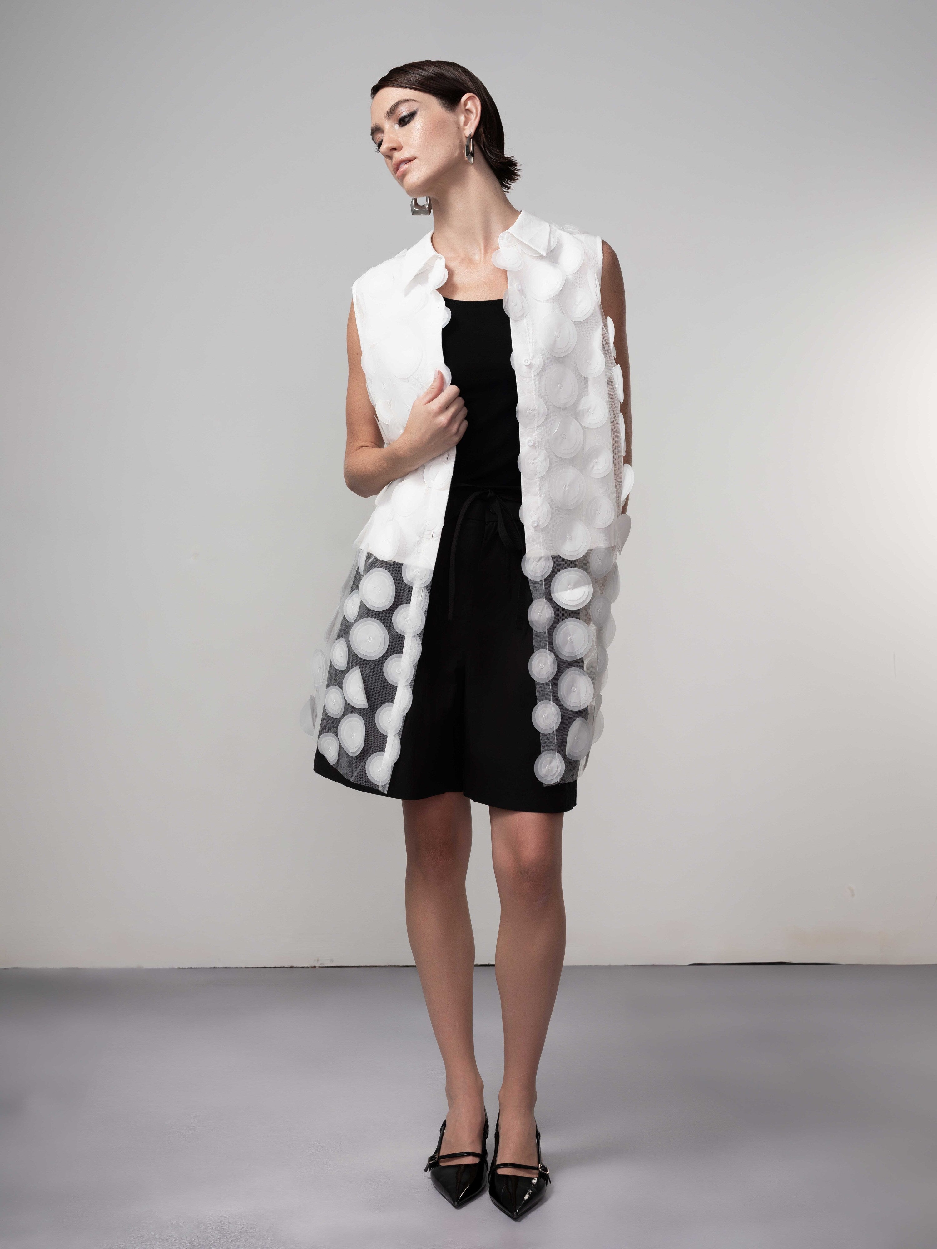 Model wearing a black sleeveless dress layered with a sheer white textured tunic vest featuring circular appliqué detailing, styled with pointed-toe heels, front view.