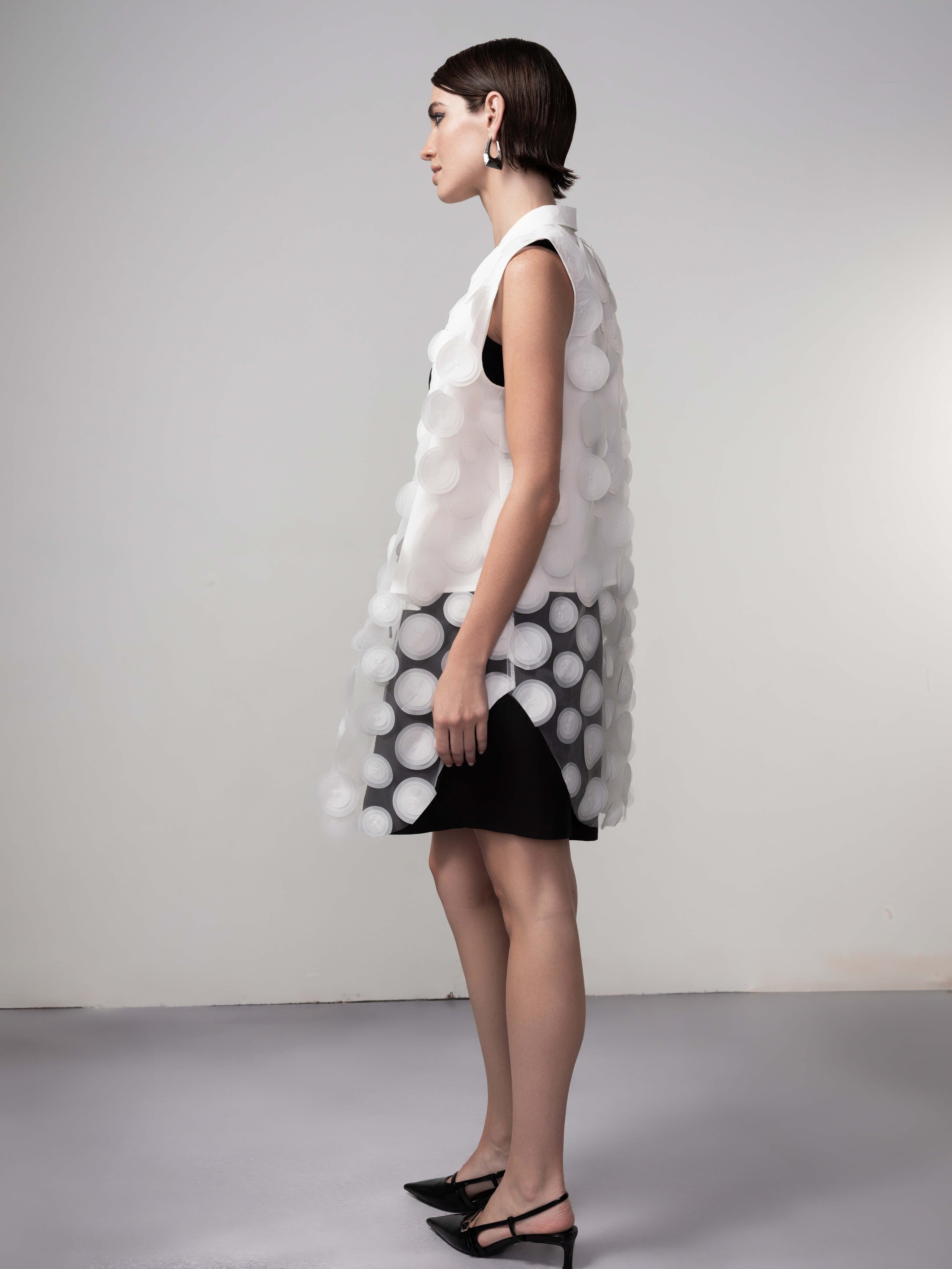 Side view of model in a black dress with a sheer white tunic vest overlay featuring raised circular texture, showing the sleeveless cut and knee-length silhouette.