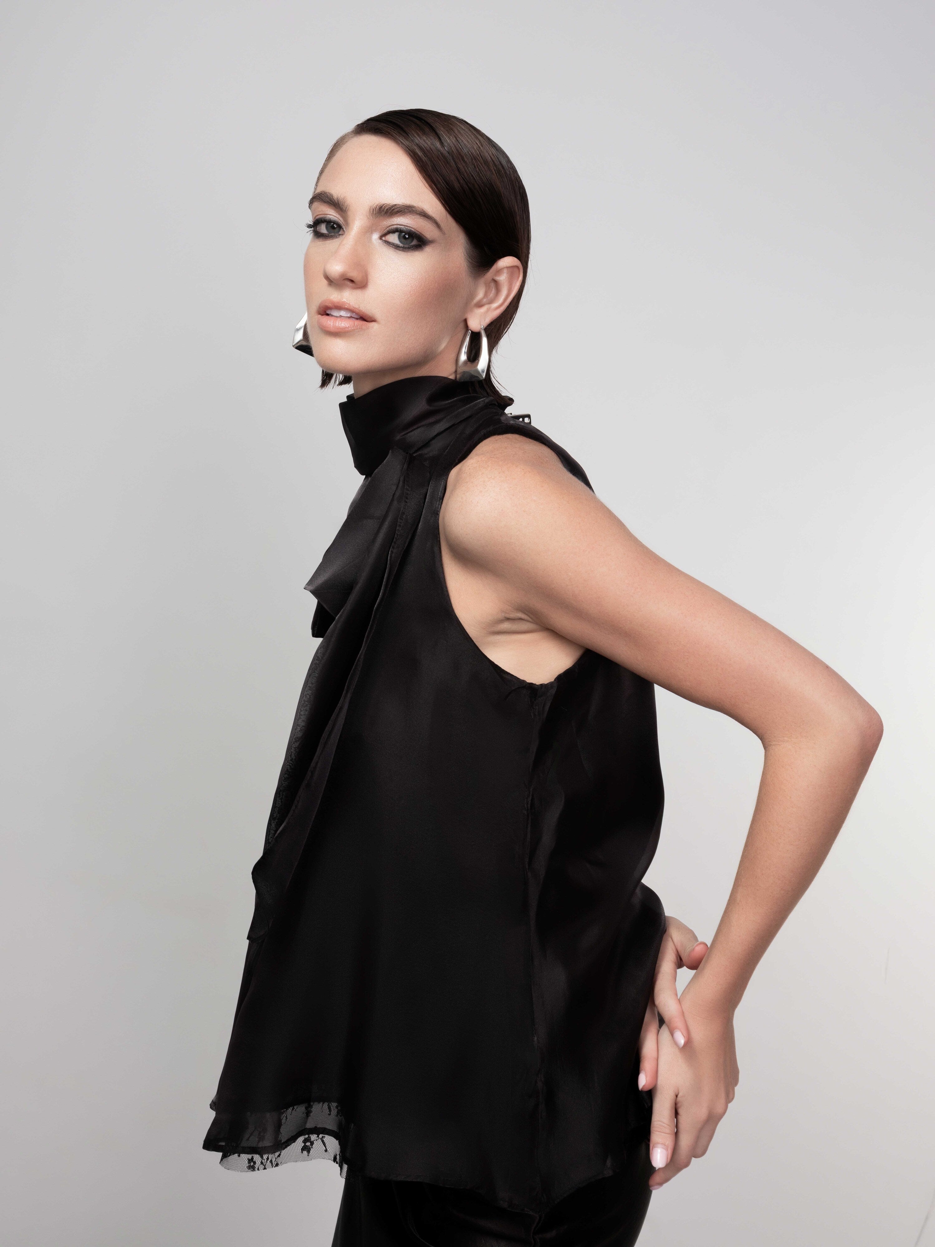 Woman wearing a black sleeveless satin blouse with tie-neck, shown from the side with a sleek, relaxed fit.
