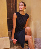 Women's sleeveless midi dress featuring elegant side ruching and a draped silhouette, styled with statement earrings while seated outdoors against a textured architectural backdrop.