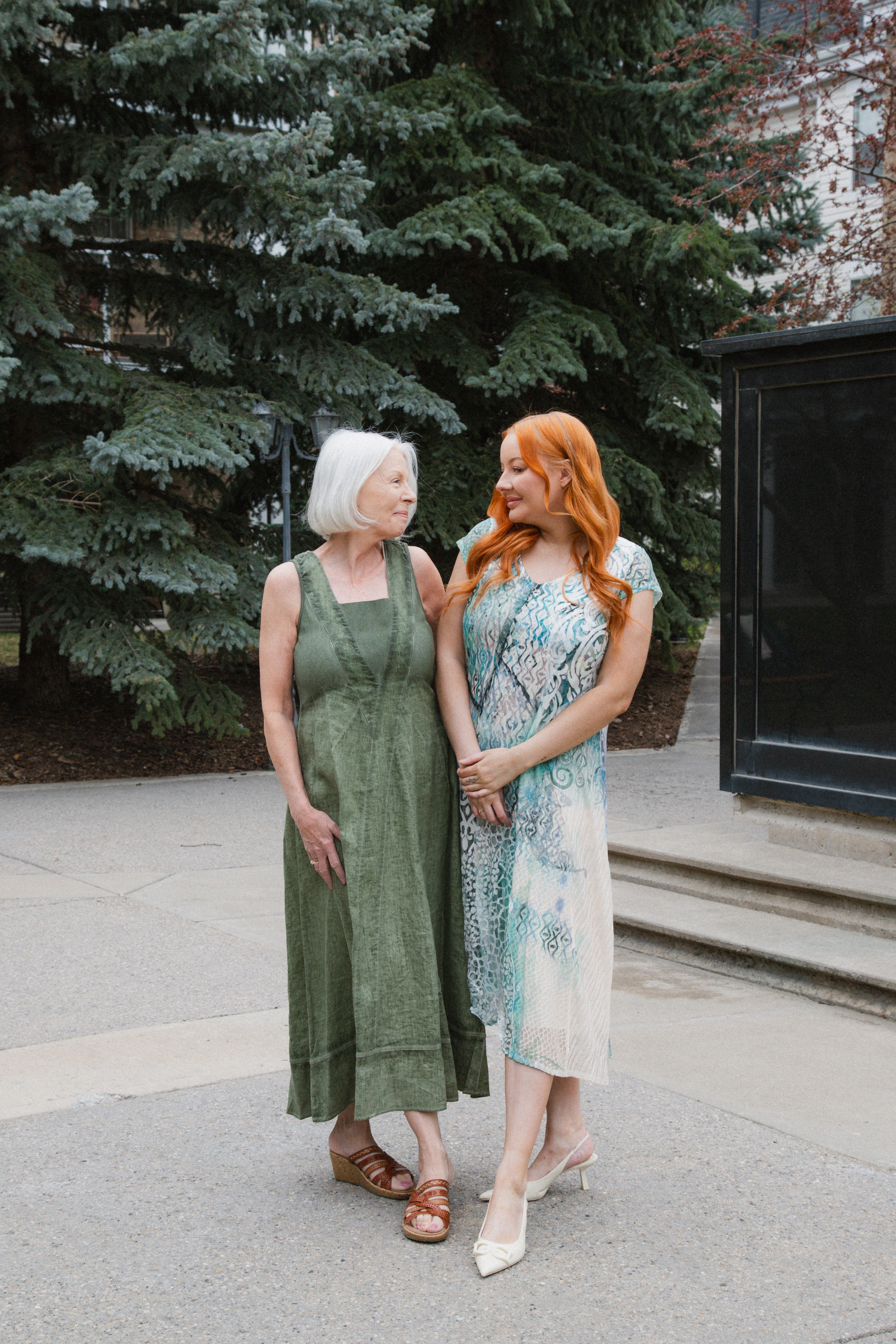 Two women standing outdoors, one with white hair and green dress, the other with red hair and floral dress, with trees and a building in the background.