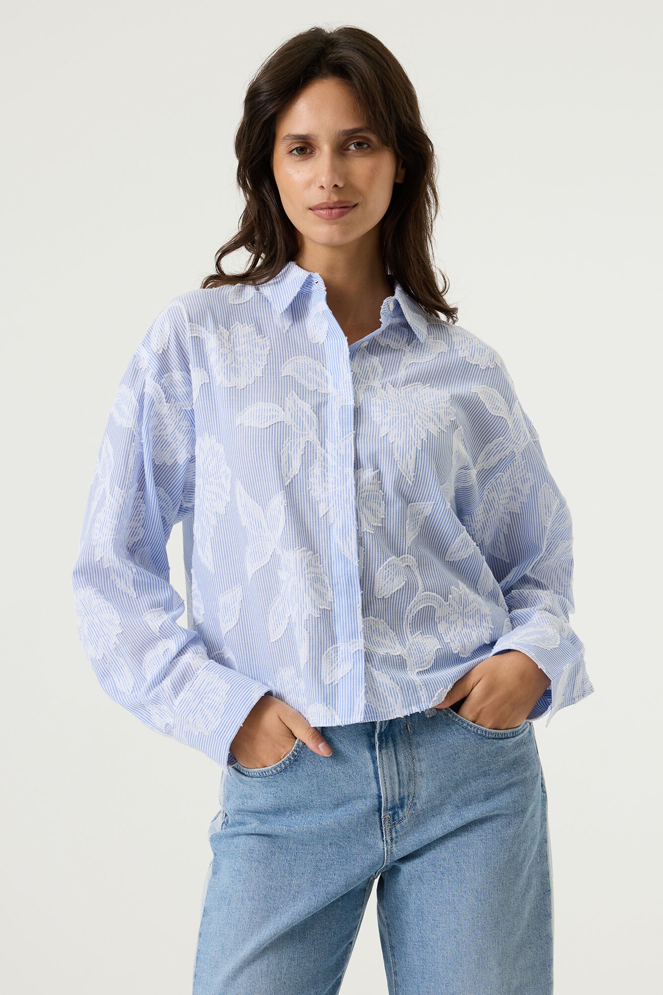 Women's light blue striped cotton blouse with textured floral detailing, featuring a collared neckline, button-front design, and relaxed fit. Tucked into light wash wide-leg jeans for an effortless everyday look.