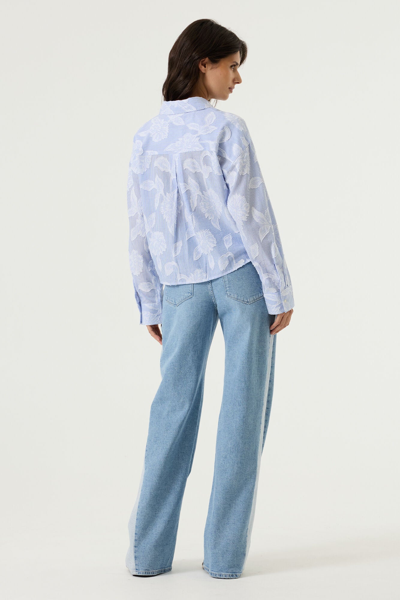 Back view of a women's light blue striped cotton blouse with textured floral detailing, showcasing a relaxed silhouette, long sleeves, and a slightly cropped hem. Styled with light wash wide-leg jeans.