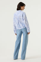 Back view of a women's light blue striped cotton blouse with textured floral detailing, showcasing a relaxed silhouette, long sleeves, and a slightly cropped hem. Styled with light wash wide-leg jeans.