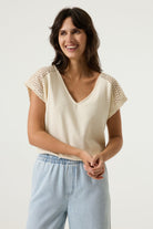 Women's cream V-neck top with crochet lace cap sleeves and a relaxed fit, styled with light wash wide-leg jeans. Front view showing the textured fabric and feminine silhouette.