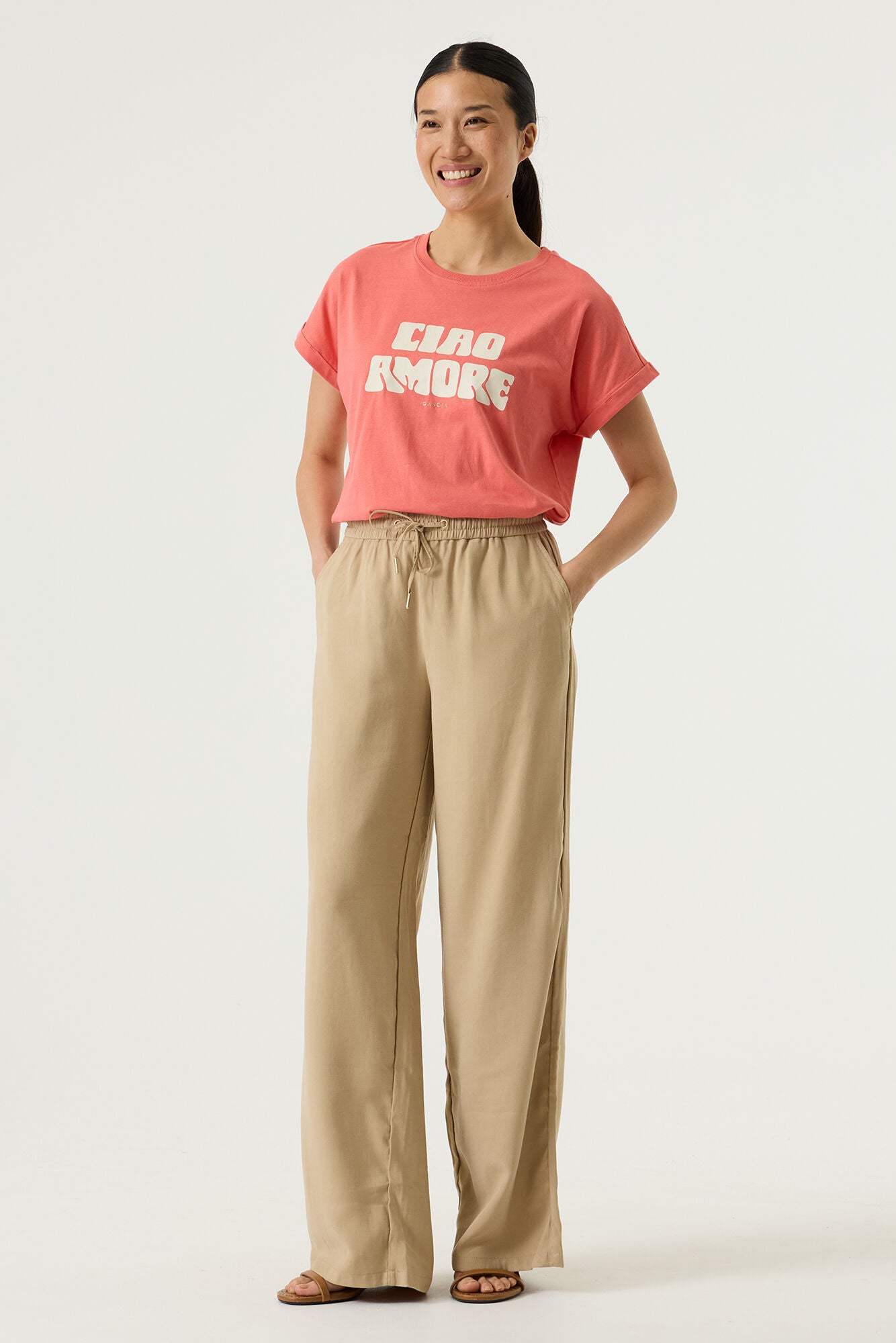 Woman wearing a coral relaxed fit graphic tee with “Ciao Amore” text in light beige, styled with beige wide-leg drawstring pants