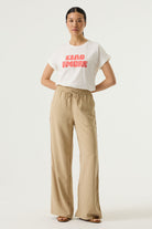 Woman wearing a white relaxed fit graphic tee with “Ciao Amore” text in pink, paired with beige wide-leg drawstring pants and flat sandals
