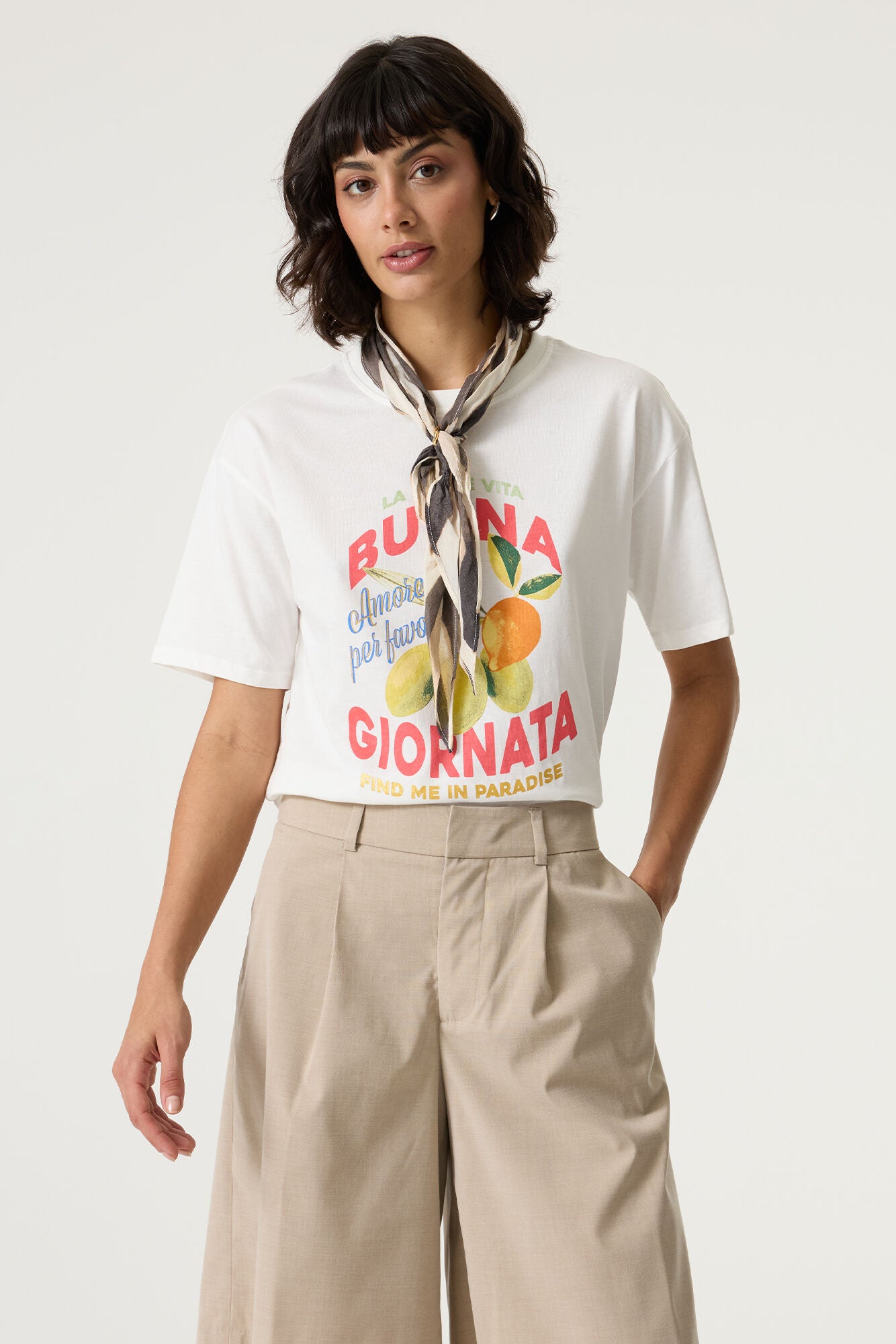 Front view of a women’s white cotton graphic t-shirt with red and blue text and a citrus fruit print, styled with beige tailored shorts and a neck scarf.