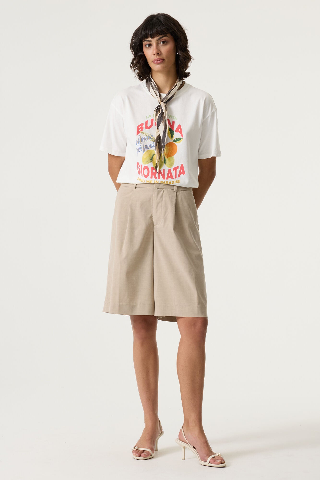 Front view of a women’s white cotton graphic t-shirt with red and blue text and a citrus fruit print, styled with beige tailored shorts and a neck scarf.