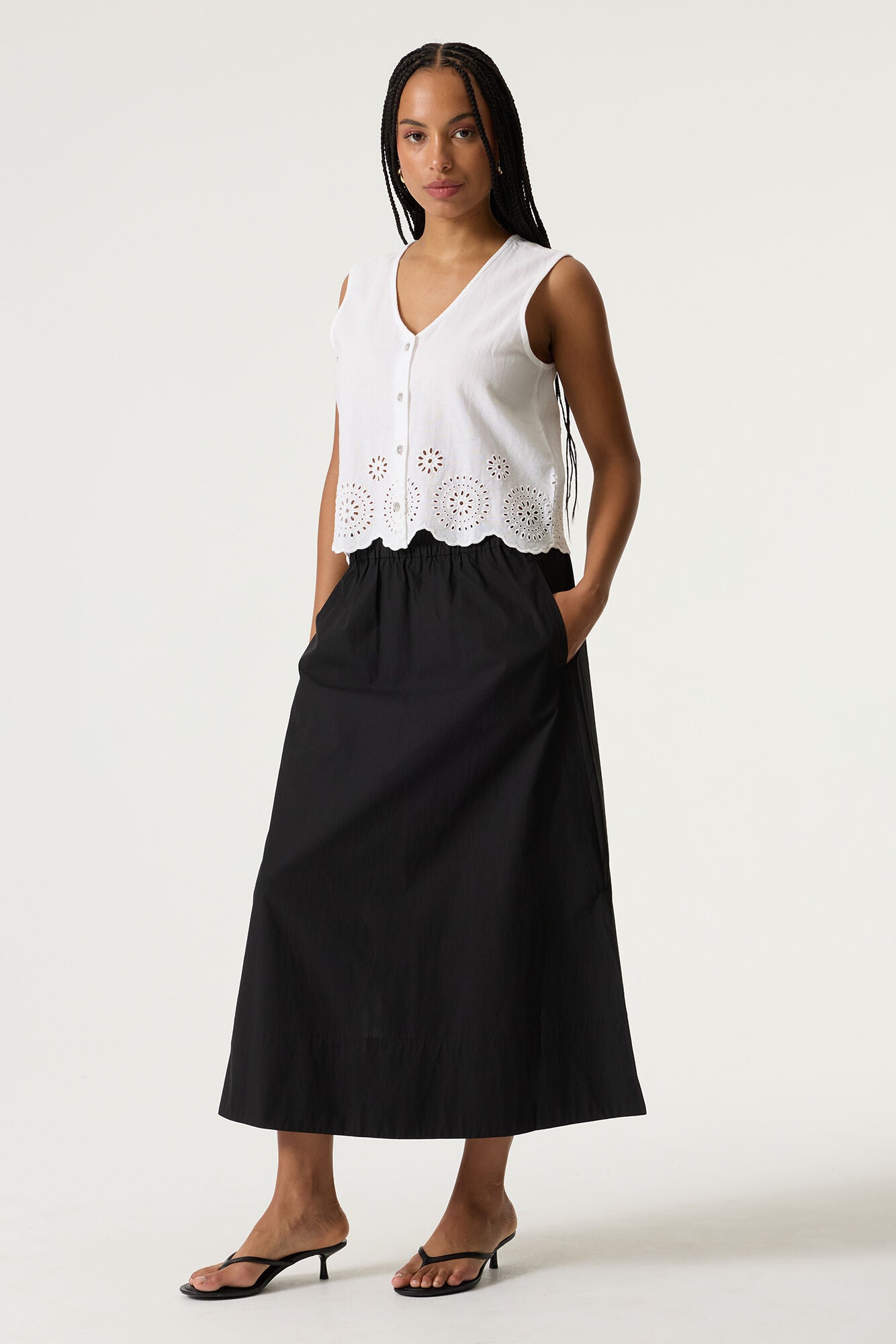Women's white sleeveless cotton-linen blouse with eyelet embroidered scalloped hem and button-front detail. Styled with a midi skirt and shown in a relaxed front-facing pose.