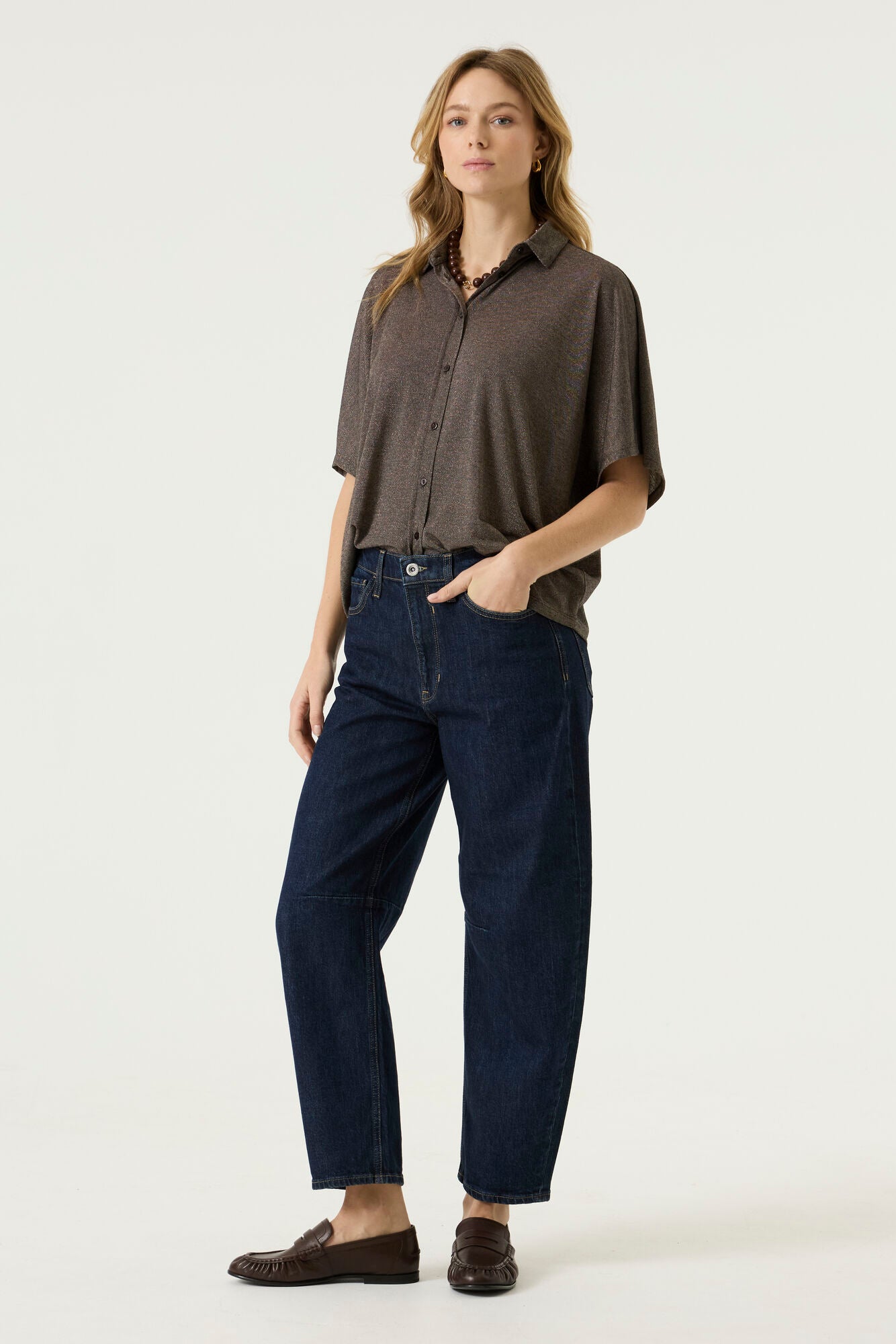 Front view of a woman wearing a brown relaxed-fit short-sleeve button-front blouse with a pointed collar, paired with dark blue jeans.