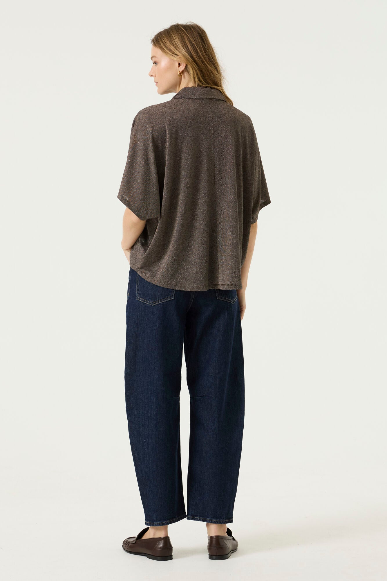 Back view of a woman wearing a brown oversized short-sleeve blouse with a relaxed drape, paired with dark blue wide-leg jeans and brown loafers.