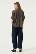 Back view of a woman wearing a brown oversized short-sleeve blouse with a relaxed drape, paired with dark blue wide-leg jeans and brown loafers.