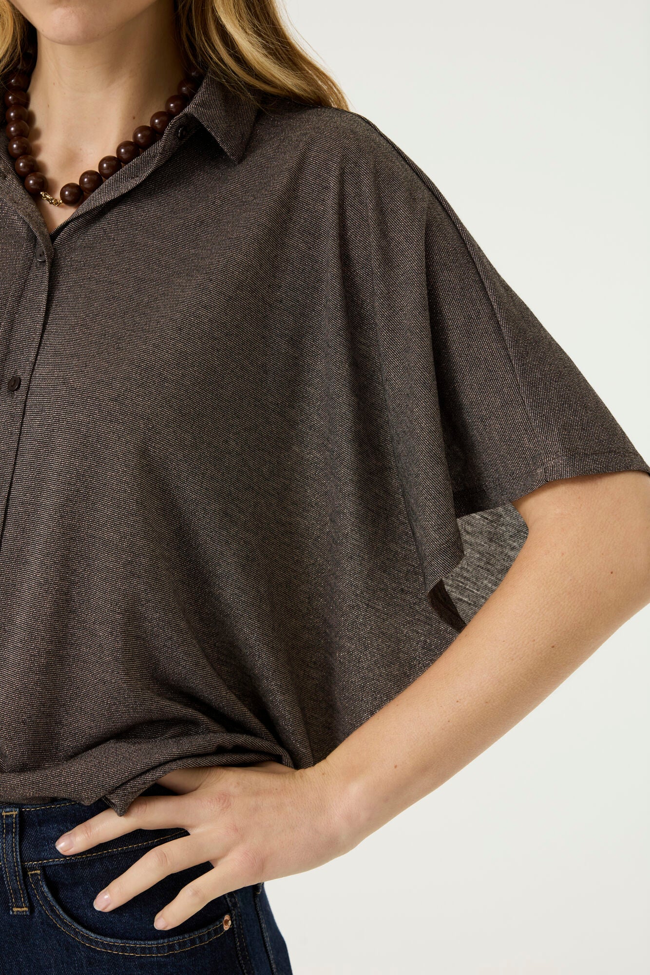 Close-up of a woman wearing a brown short-sleeve button-front blouse with a relaxed fit, pointed collar, and subtle textured fabric, paired with dark blue jeans.