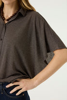 Close-up of a woman wearing a brown short-sleeve button-front blouse with a relaxed fit, pointed collar, and subtle textured fabric, paired with dark blue jeans.